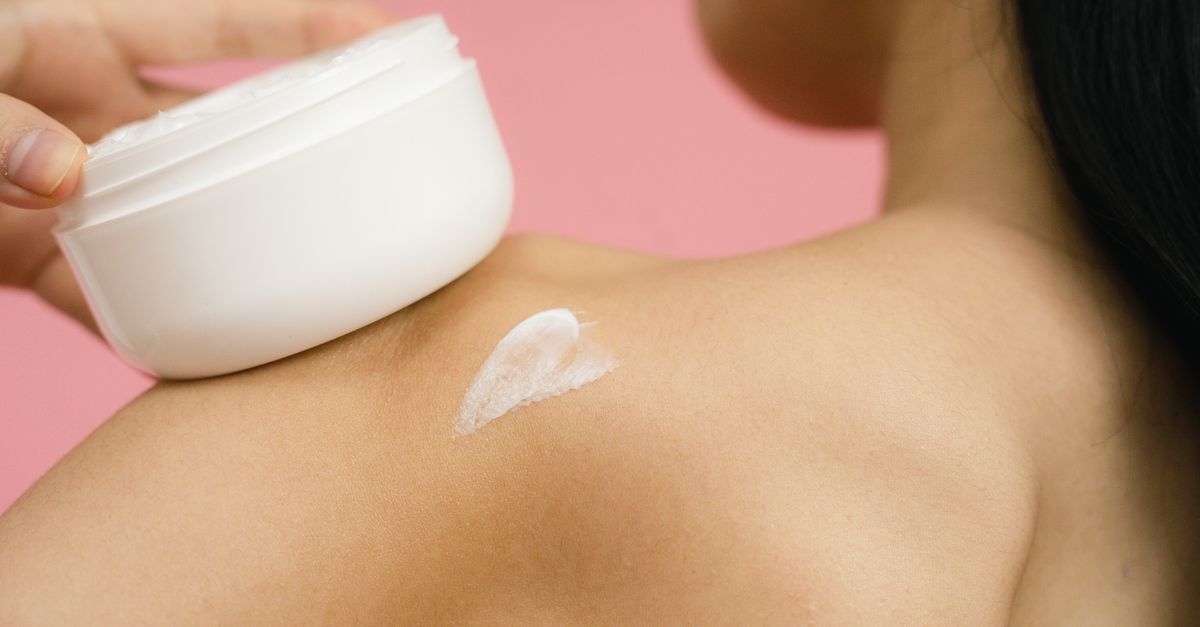 Woman applying skincare cream to shoulder against a pink background.