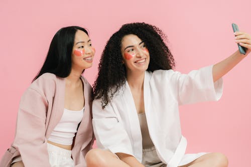 Two women with cosmetic pads sitting together on floor and posing for selfie