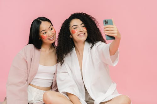 Two women with cosmetic pads sitting on floor and smiling for selfie