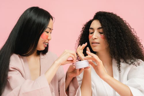 Two women picking cosmetic product up from white round box
