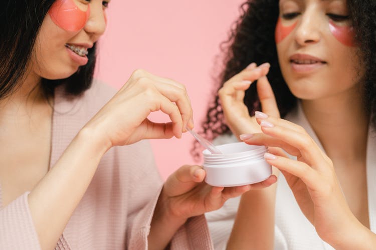 Two Women Picking Up Cosmetic On Spatula And Finger From Round Small Box