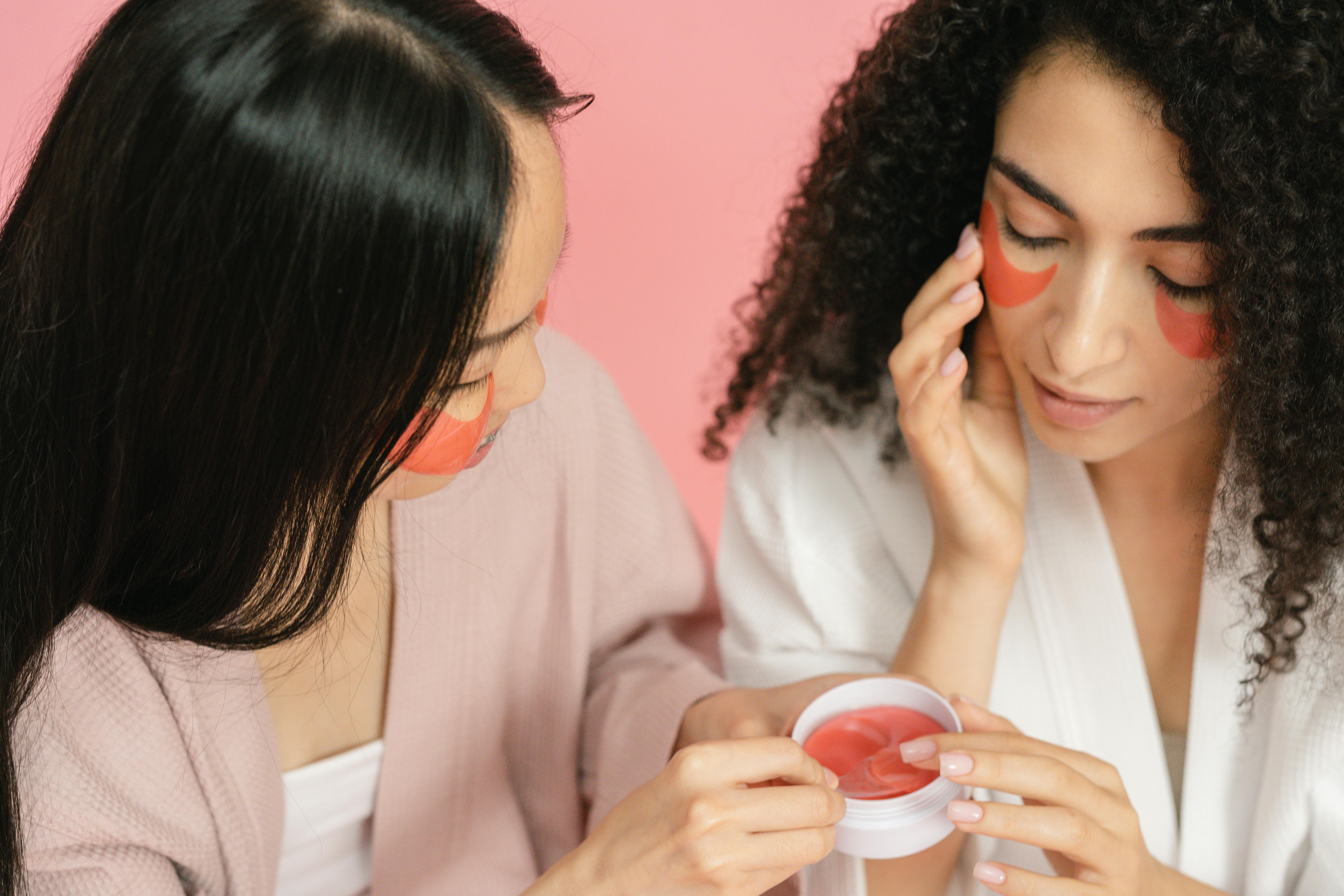 two women using red cosmetic pads and putting them on under eyes