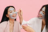 Woman with cosmetic mask on her face putting gray clay on other womans face