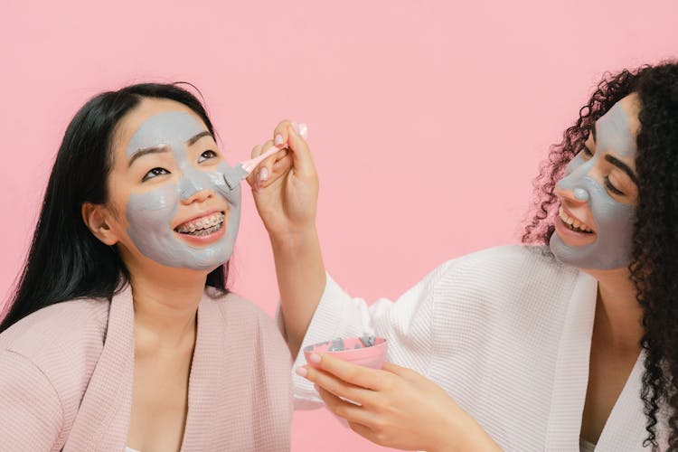 Woman With Cosmetic Mask On Her Face Putting Gray Clay On Other Womans Face