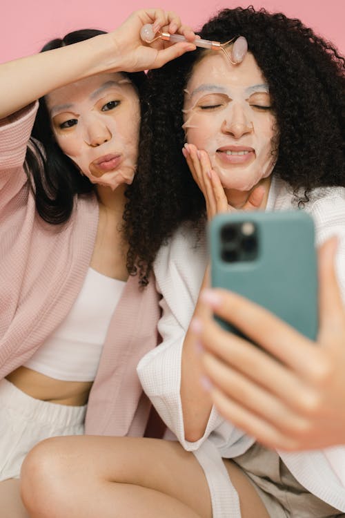 Two women in bathrobes with cosmetic wrapping masks on faces making selfie