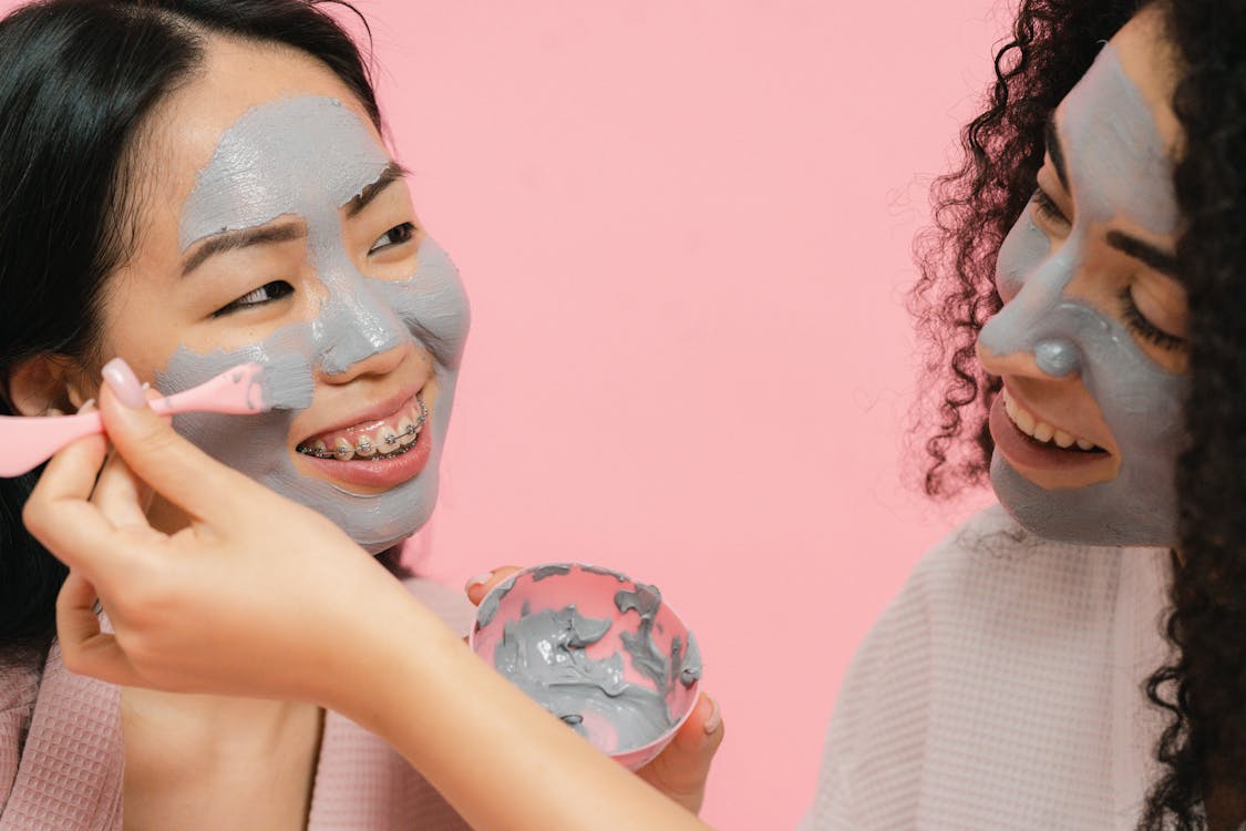 Woman with cosmetic mask on her face putting on gray clay on face of other woman