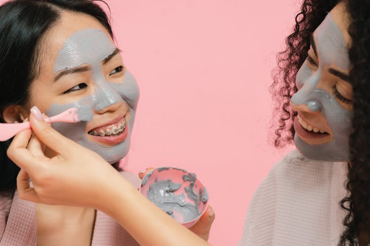 Woman With Cosmetic Mask On Her Face Putting On Gray Clay On Face Of Other Woman