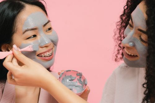 Free Two women applying gray clay facial masks, smiling and enjoying a relaxing skincare session. Stock Photo