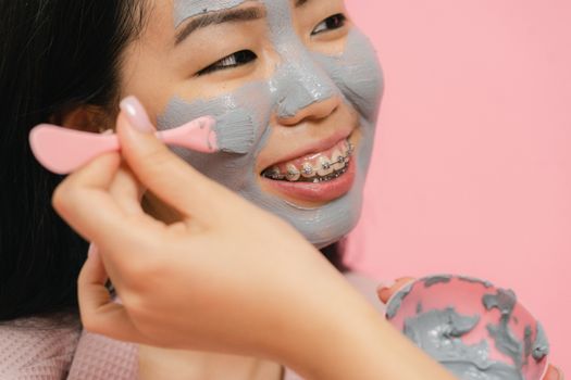 Close-up of a woman with braces applying a clay face mask, smiling cheerfully against a pink background.