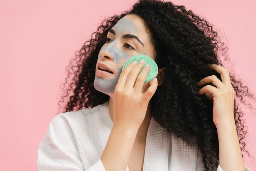 Adult woman with curly hair applying a facial mask using a sponge in a studio setting.