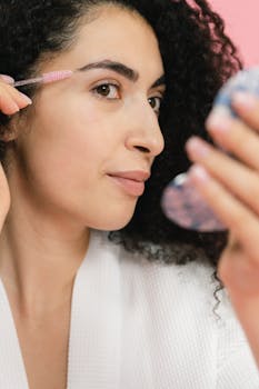 A woman with curly hair using a brush to groom her eyebrows in a reflective mirror indoors.