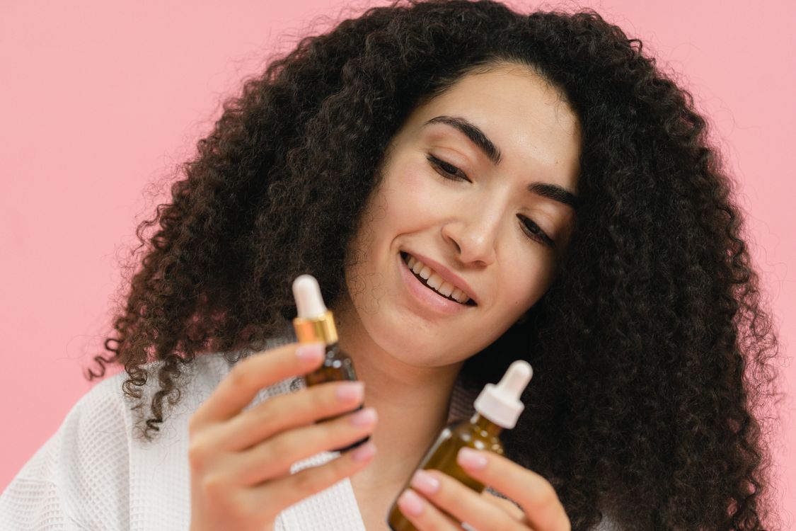 Free A young woman analyzes skincare bottles, set against a pink backdrop. Stock Photo