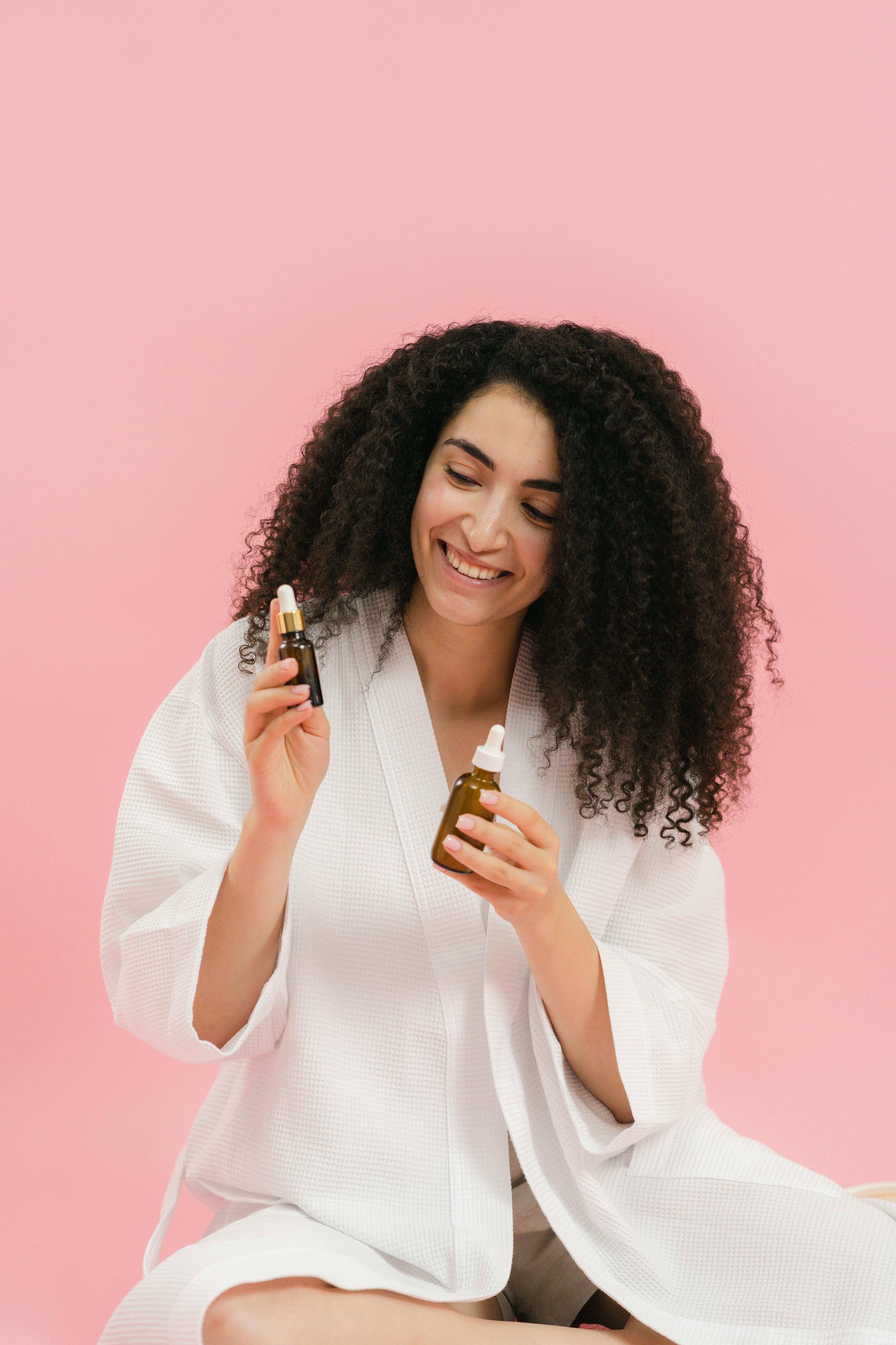 Free Woman in a bathrobe smiles while holding facial serums against a pink background. Stock Photo
