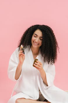 Woman in a bathrobe smiles while holding facial serums against a pink background.