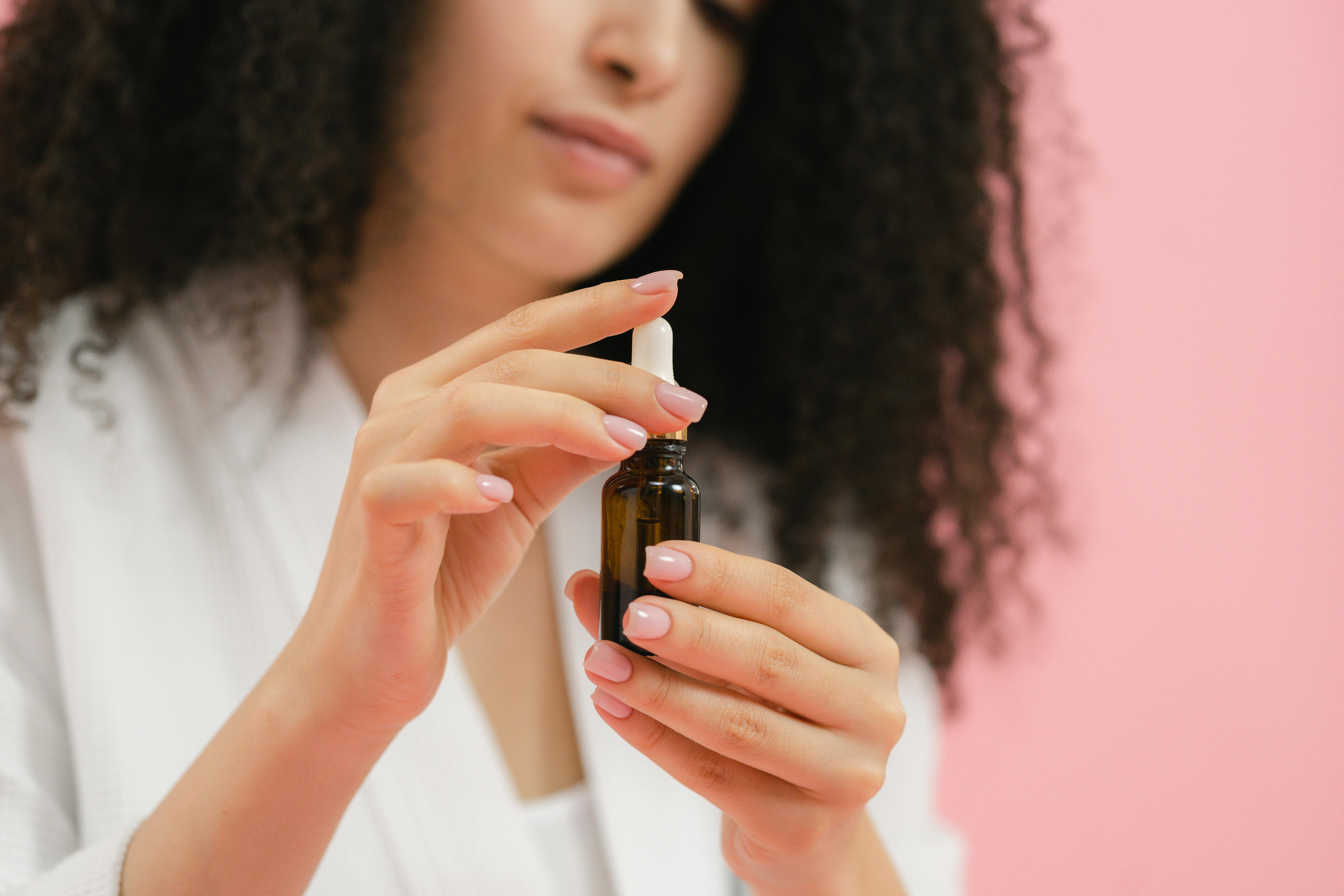 Close-up of woman holding a glass dropper bottle for skincare against a pink background.