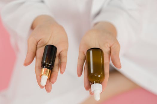 A close-up of hands holding two glass skincare bottles on a soft pink background.
