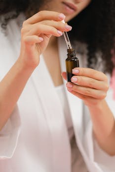 A woman in a white robe uses a dropper to apply skincare serum from a glass bottle.