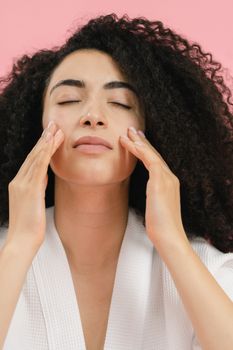 Woman with curly hair and closed eyes enjoying a skincare routine against a pink background.