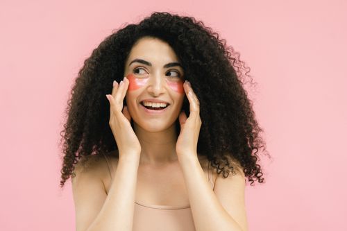 Free Happy woman with curly hair applying pink under eye patches on a pastel pink background. Stock Photo
