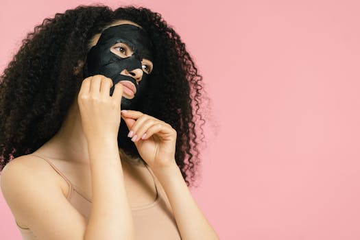A woman with curly hair applies a black mask on pink backdrop, emphasizing skincare.