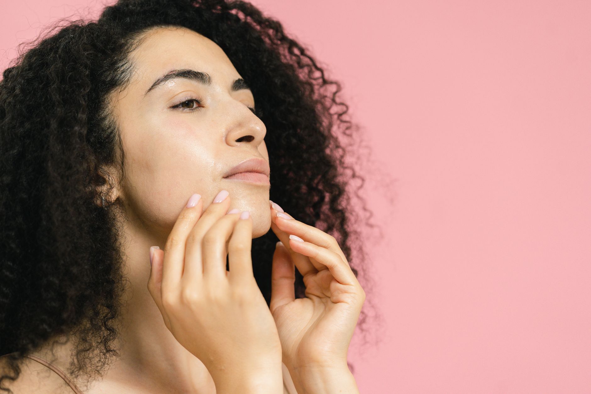 Anti ageing protocol — A woman with curly hair touches her face, set against a pastel pink background.