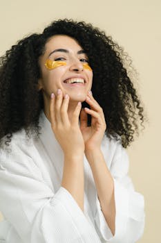 Radiant woman with curly hair applying skincare under eye patches indoors.