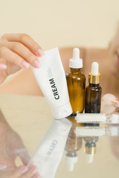 A hand reaching for a cream tube among skincare bottles on a reflective surface.