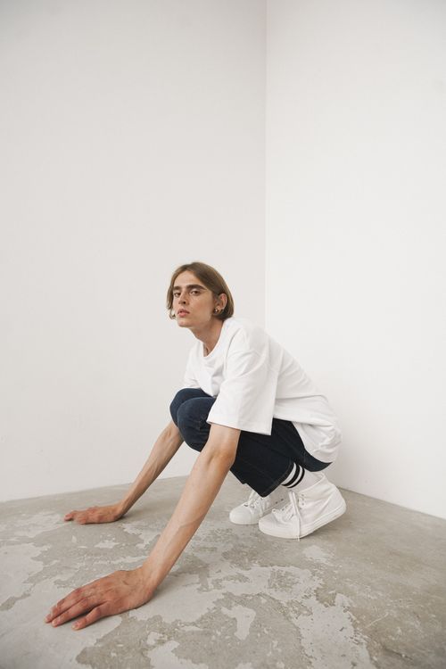Free A young man wearing casual attire crouching in a minimalist room with white walls and floor. Stock Photo