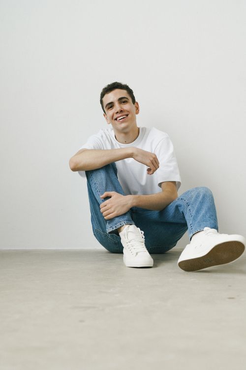 Free Young man sitting casually on the floor, smiling, wearing denim jeans and a white T-shirt against a clean white background. Stock Photo