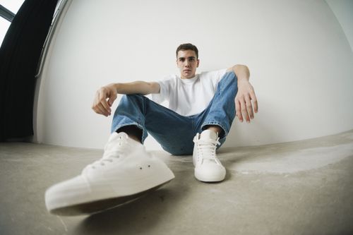 Free Young man in casual attire sitting in a minimalist studio, showcasing modern fashion style. Stock Photo