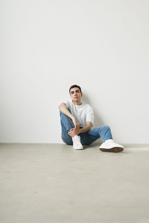 Free A young man sits casually against a white wall, wearing a white shirt and jeans. Stock Photo