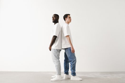 Two men standing back to back wearing white T-shirts and denim pants against a white background. Represents diversity and style.