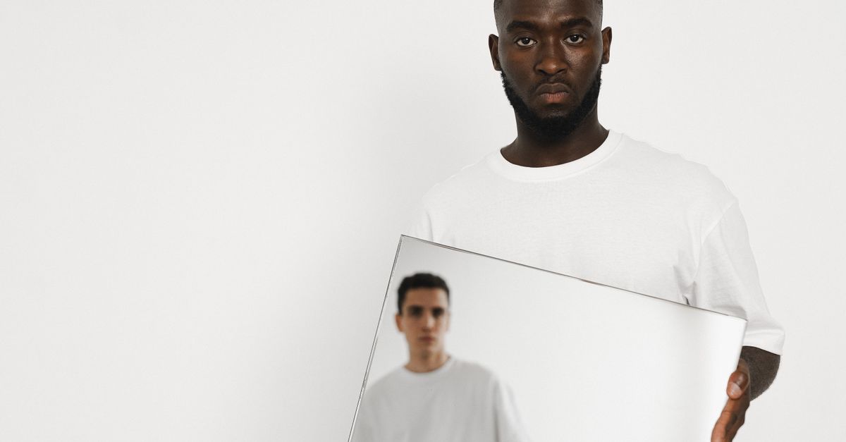 A man in a white shirt holding a mirror reflecting another person indoors