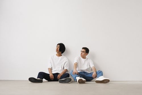 Free Two young men sitting casually in a minimalist indoor environment, wearing white shirts and jeans. Stock Photo