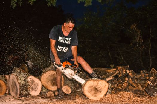 A man using a chainsaw to cut logs outdoors at night, surrounded by wood.