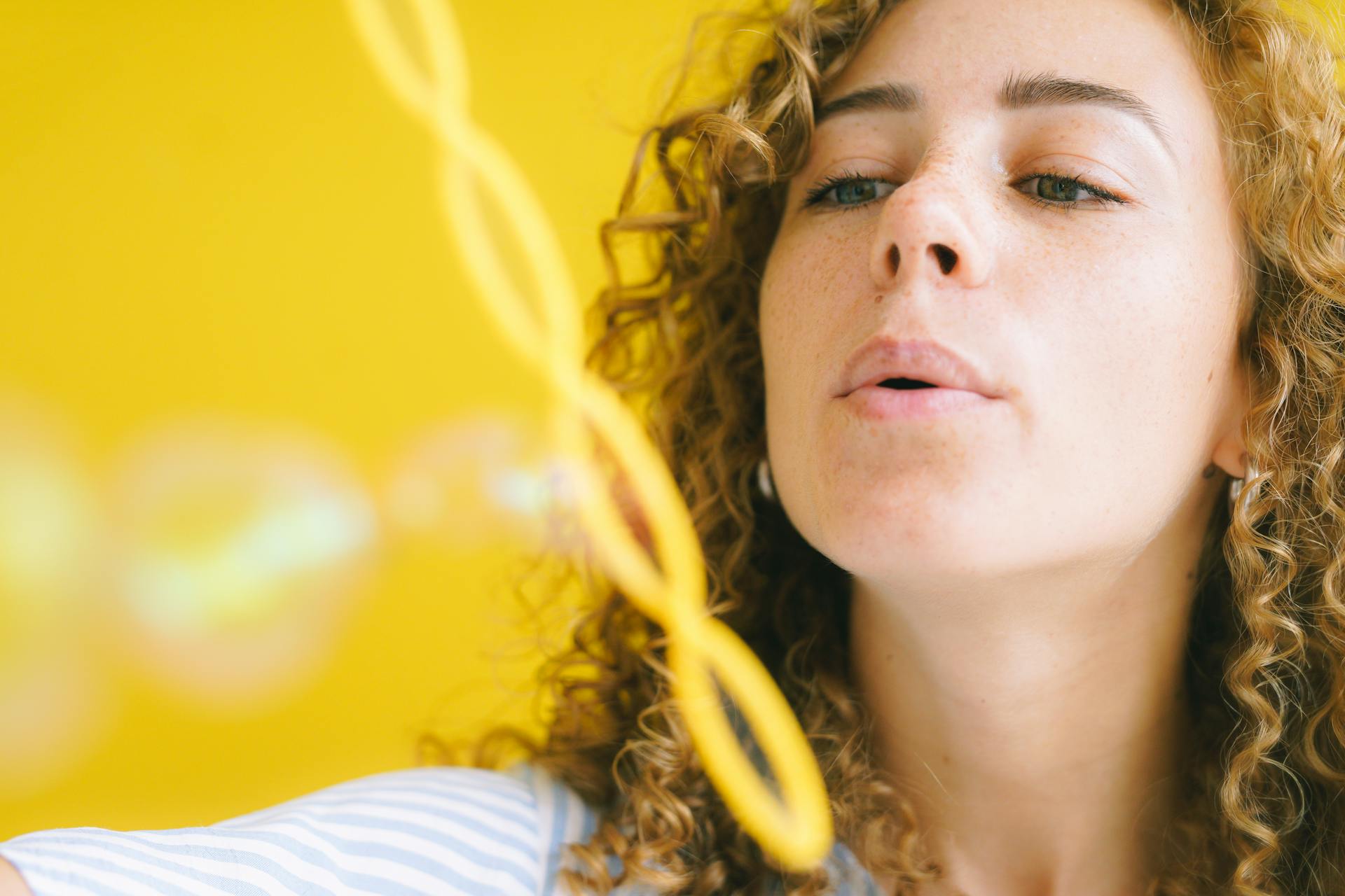 Close-up of a woman blowing bubbles with a playful expression against a bright yellow background.