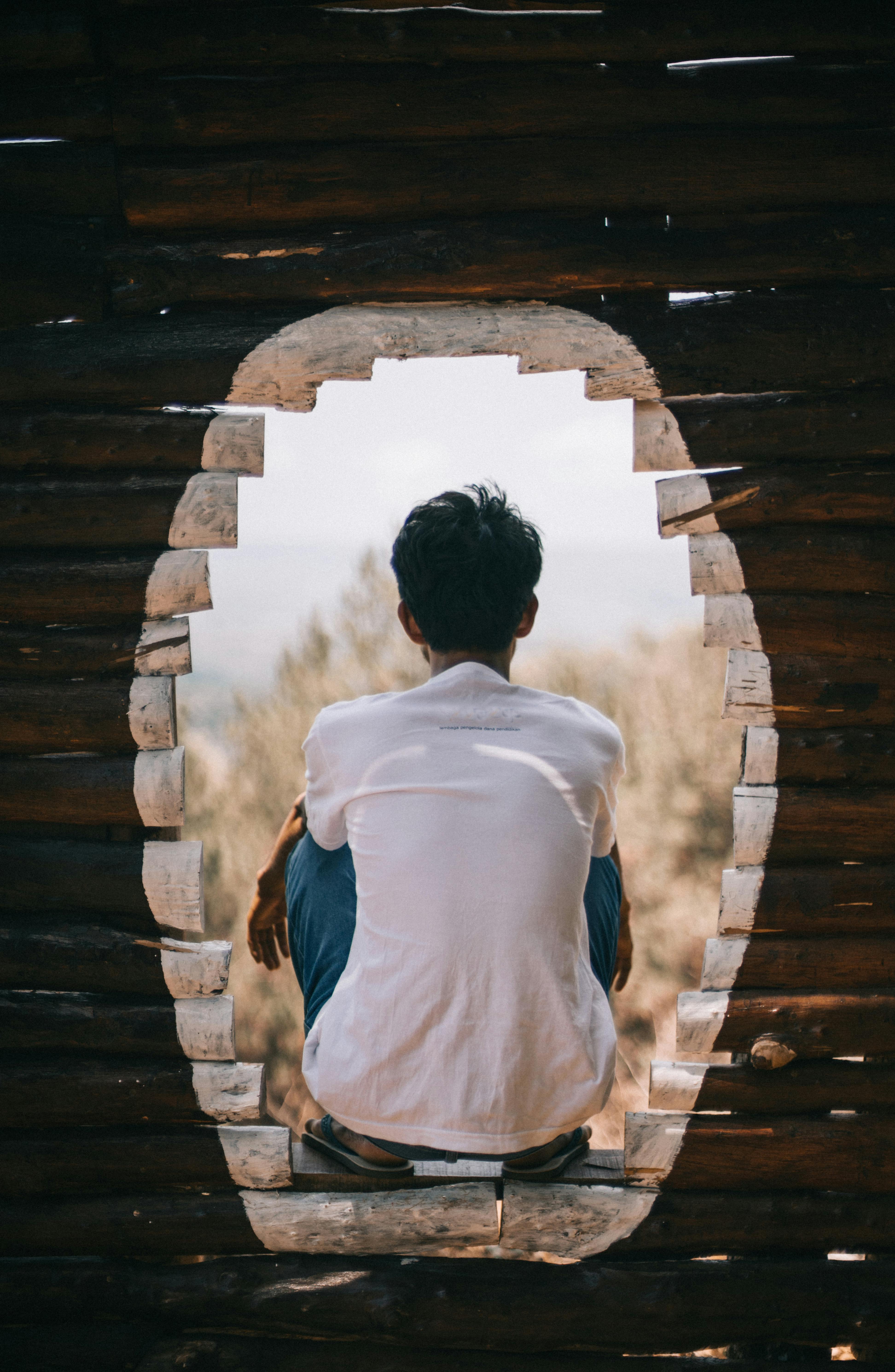 A contemplative moment captured as a man sits in a round window of a rustic wooden structure, amidst nature.