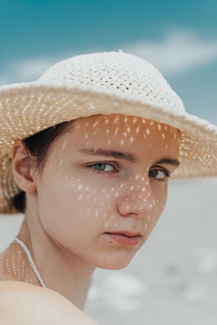 A young woman outdoors under dappled sunlight, wearing a straw hat, with an intense gaze.