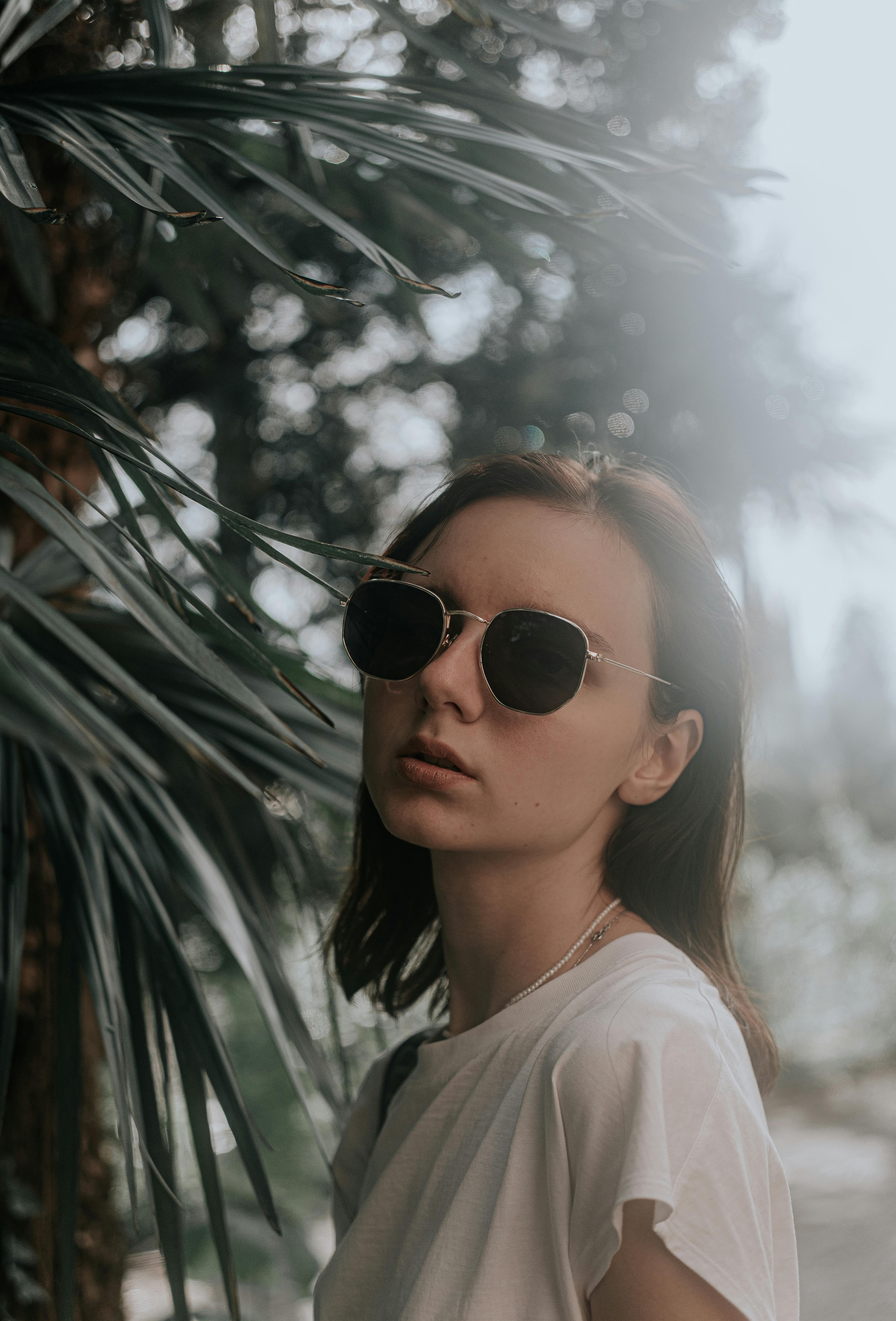 A woman wearing sunglasses, standing outdoors surrounded by tropical foliage, exuding a relaxed vibe.