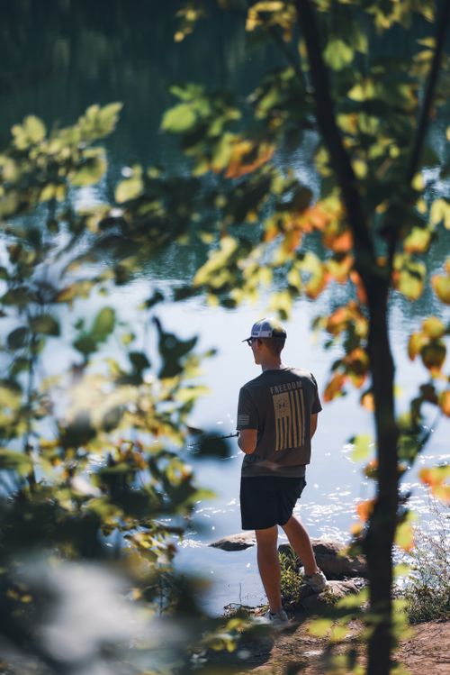 Free Man in casual attire standing by a peaceful lakeside surrounded by nature. Stock Photo