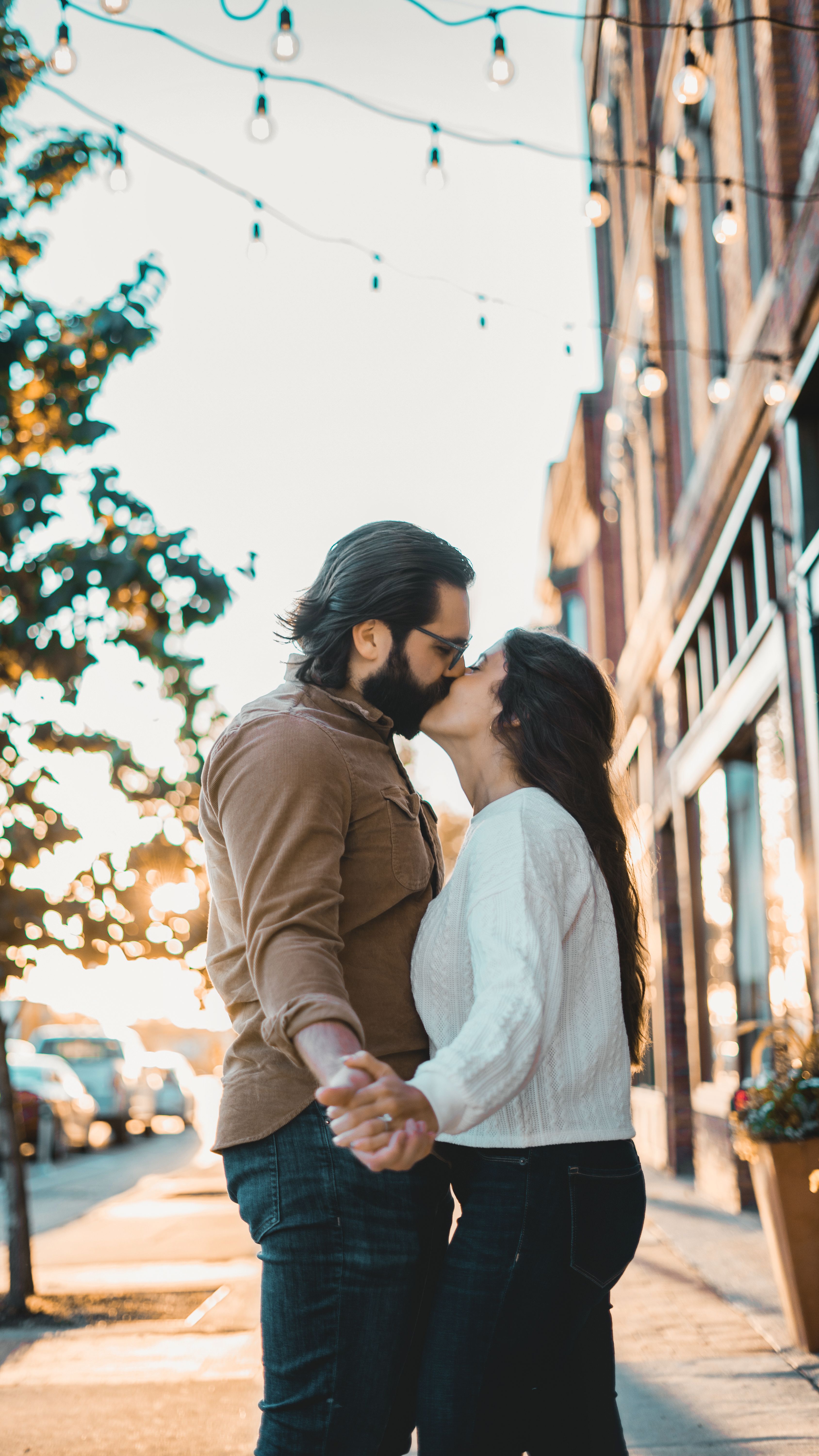 A romantic couple shares a kiss on a city street at sunset, holding hands warmly.