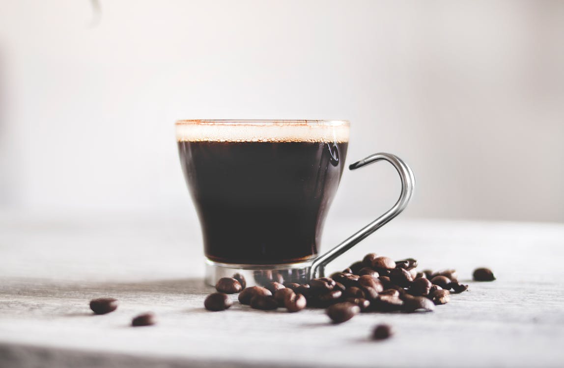 Free Aromatic black coffee in glass cup with metal handle and scattered coffee beans on light wooden table against blurred background Stock Photo