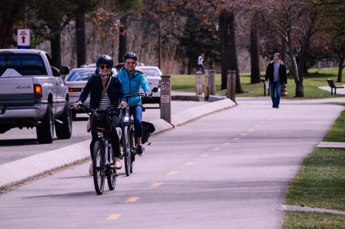 Free Two women cycling on an urban bike path in a park with vehicles nearby. Stock Photo
