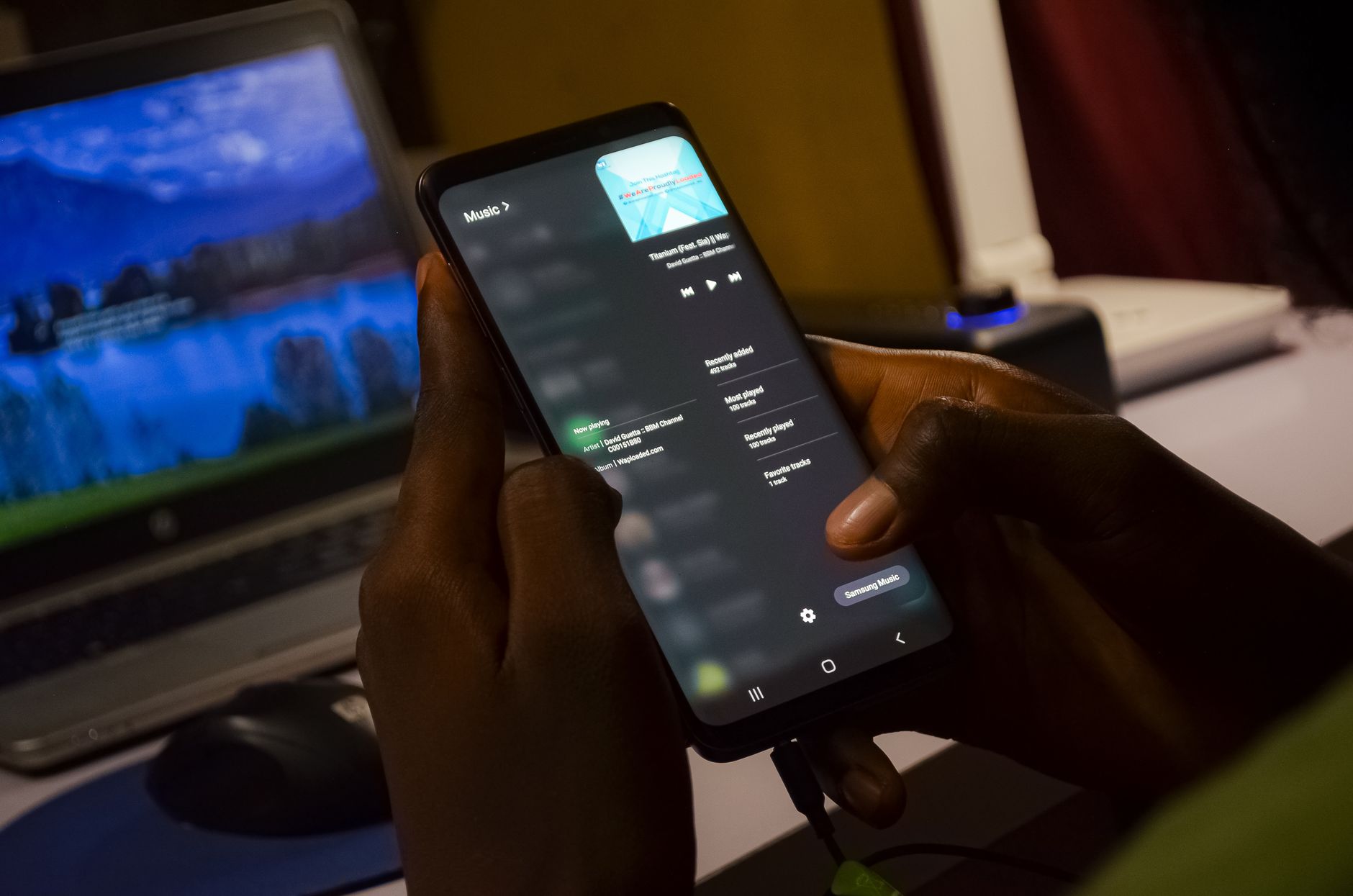 Person using smartphone with music app, indoors with laptop in background.