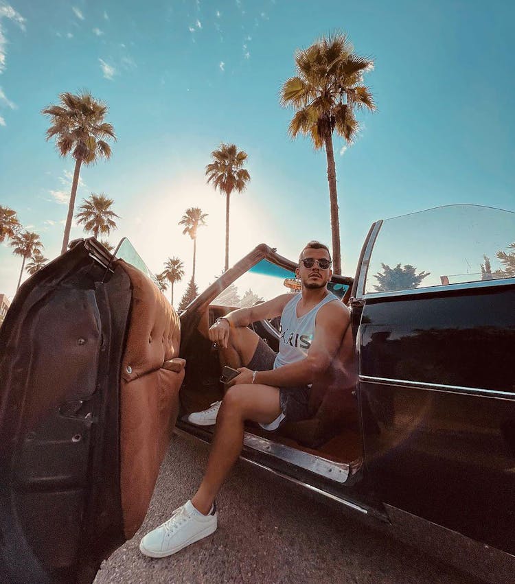 A Man In White Tank Top And Sunglasses Sitting On The Car