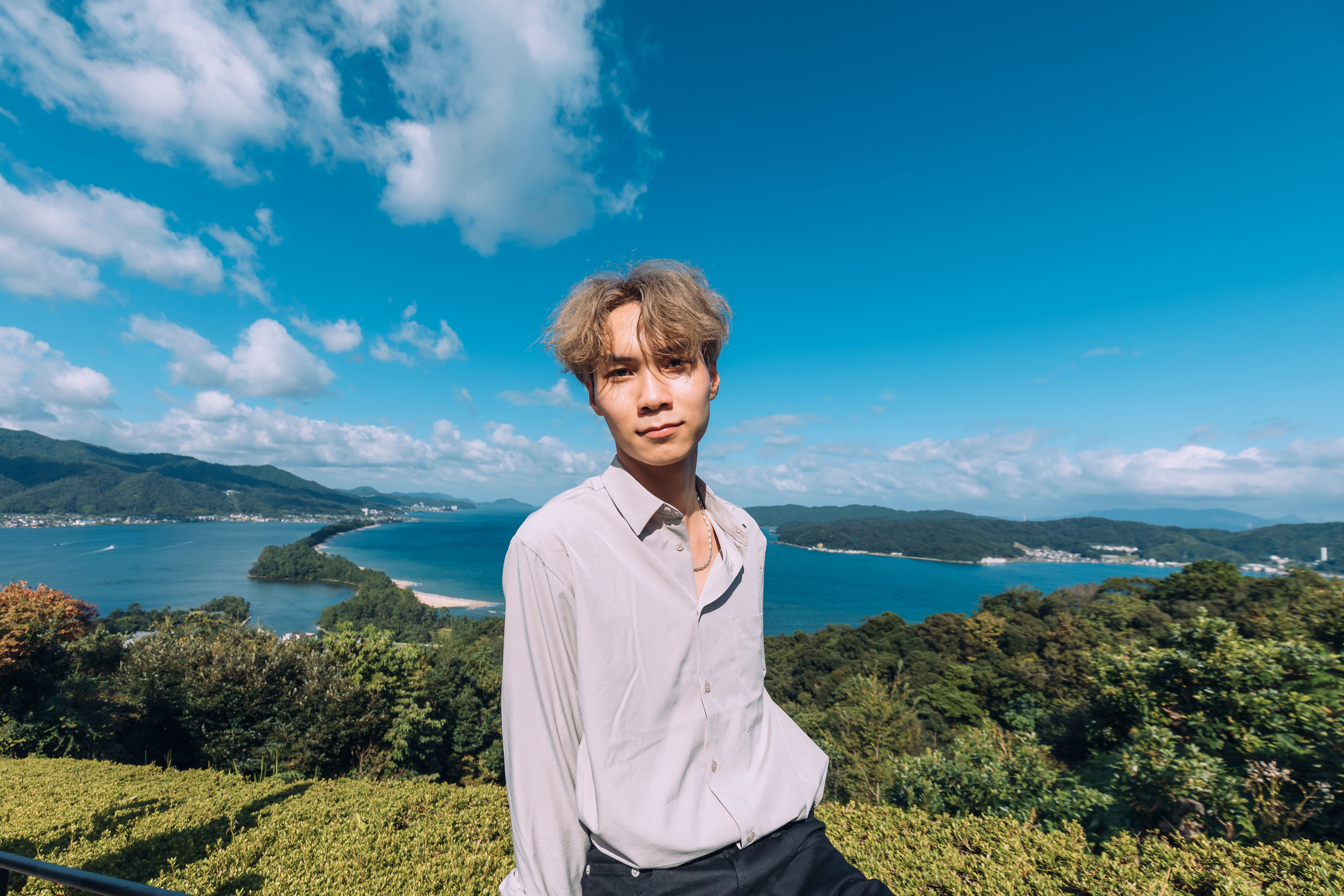 A stylish young man poses confidently on a beautiful scenic coastline, with clear blue skies and lush greenery.