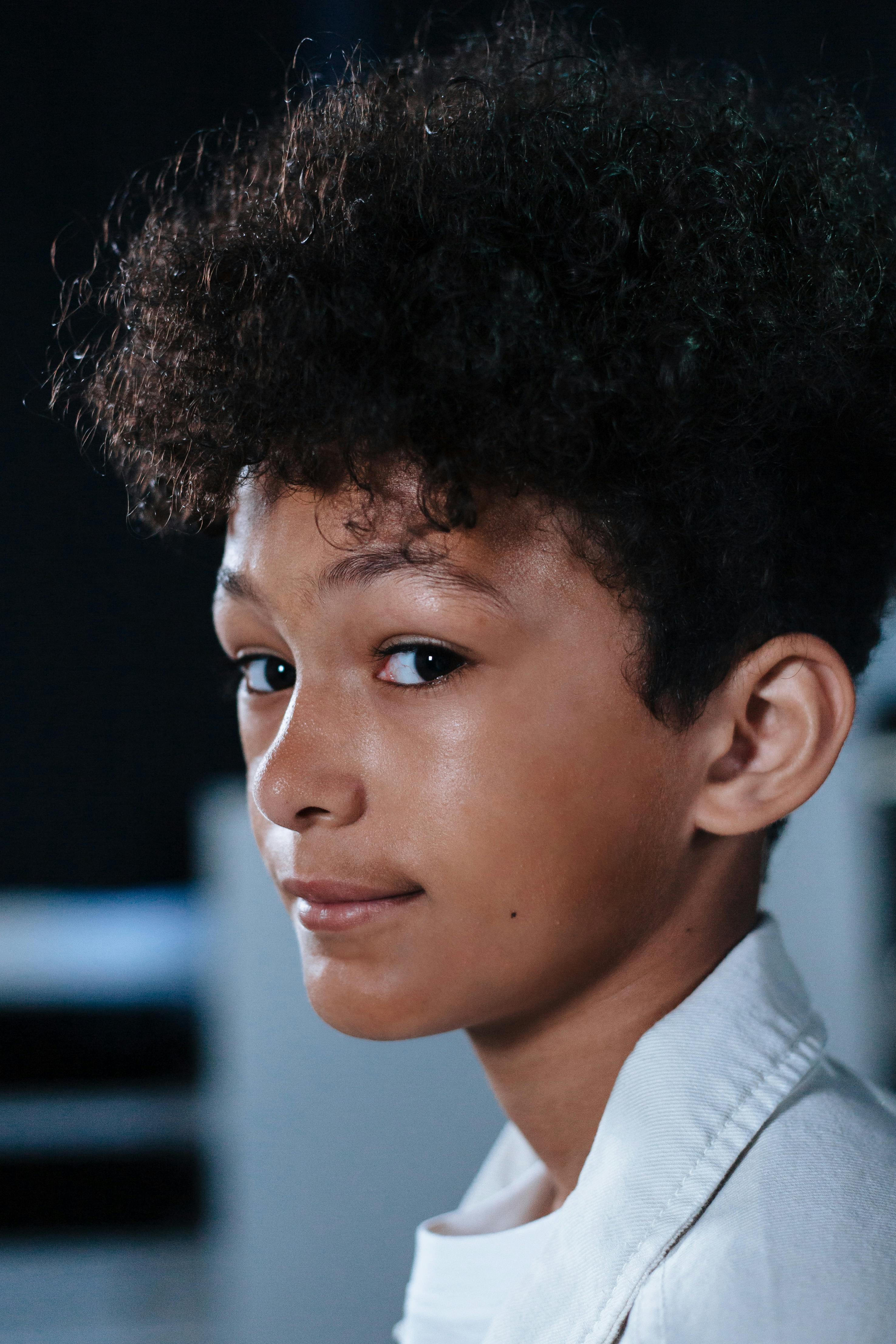 Close-up portrait of a smiling teenager with curly hair and a white shirt, looking sideways.