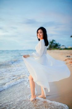 A smiling woman in a flowing white dress strolls along the vibrant beach at sunset.
