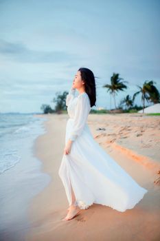 A woman in a white dress strolls along a tropical beach at dusk, feeling the sea breeze.