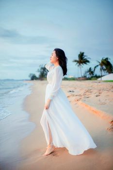 Elegant Asian woman in flowing white dress on a tranquil beach at twilight.